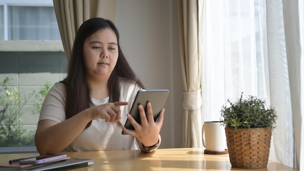 Junge mollige Frau sitzt in der Küche am Fenster und benutzt ein digitales Tablet