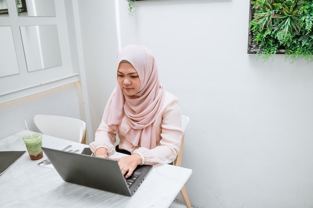 Junge moslemische Frau, die schwarzen Laptop verwendet. Junge asiatische Geschäftsfrau, die in der Kaffeestube sitzt und an Laptop arbeitet.