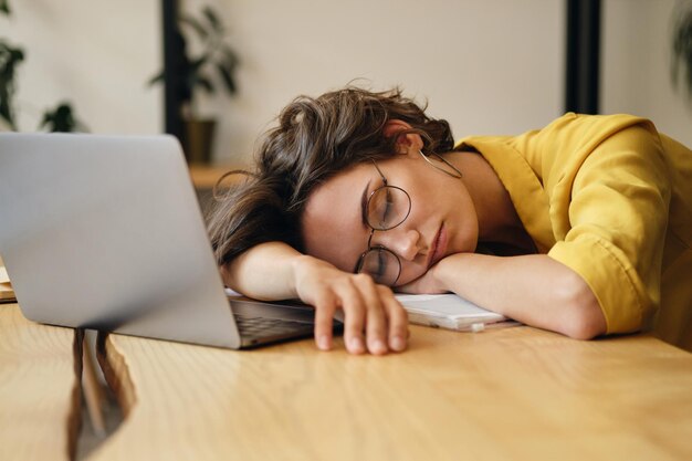 Junge müde Frau mit Brille, die allein am Arbeitsplatz mit Laptop auf dem Schreibtisch schläft