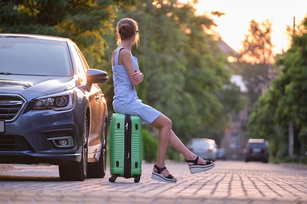 Junge müde Frau mit Koffer sitzt neben dem Auto und wartet auf jemanden. Reise- und Urlaubskonzept.