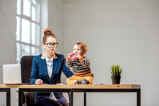 Junge Multitasking-Geschäftsfrau im Anzug, die mit Laptop und Dokumenten arbeitet, die mit ihrem kleinen Sohn im Büro sitzen?