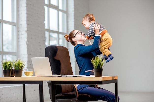 Junge Multitasking-Geschäftsfrau im Anzug, die Spaß mit ihrem kleinen Sohn im Büro hat