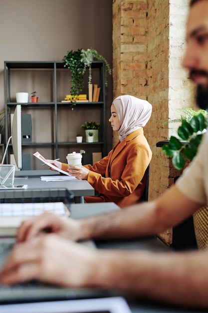 Junge muslimische Buchhalterin in Hijab und Freizeitkleidung, die durch Papier schaut