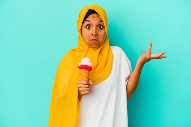Junge muslimische Frau, die ein Eis auf blauen Achselzuckenschultern und offenen Augen isst, verwirrt.