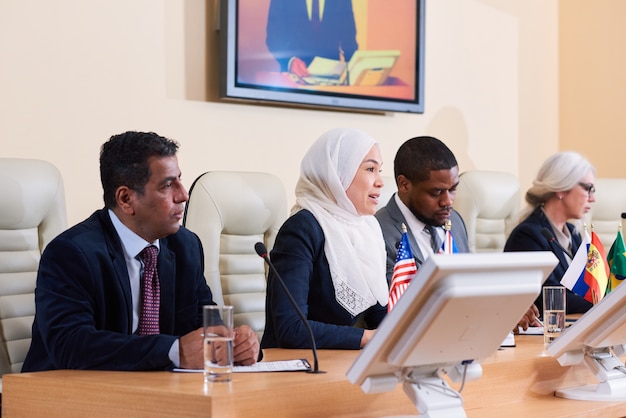 Junge muslimische Frau im weißen Hijab, die Rede auf Geschäfts- oder politischer Konferenz unter ausländischen Kollegen hält