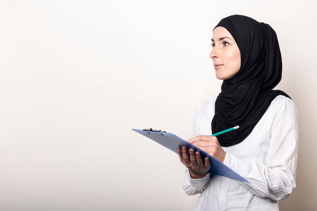 Junge muslimische Frau in einem weißen Hemd und Hijab hält eine Zwischenablage und blickt zur Seite, um das Banner für die Einstellung von Auditoren zu sehen