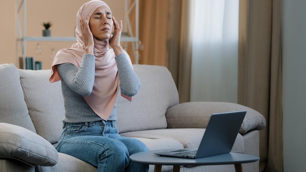 Junge muslimische frau in rosa hijab sitzt auf dem sofa und arbeitet mit laptop und hat kopfschmerzen migräne traurig