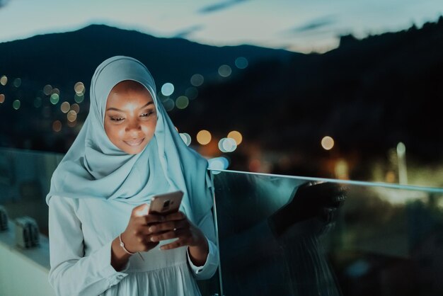 Junge muslimische Frau mit Schalschleier auf der Straße der Stadt in der Nacht, die auf einem Smartphone mit Bok SMS schreibt