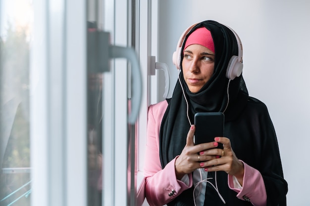 Junge muslimische Frau trägt einen Hijab mit Technologie im Innenbereich. Horizontale Seitenansicht einer arabischen Frau, die zu Hause Musik mit Smartphone neben einem Fenster hört. Technologie und Lebensstil der muslimischen Frauen.