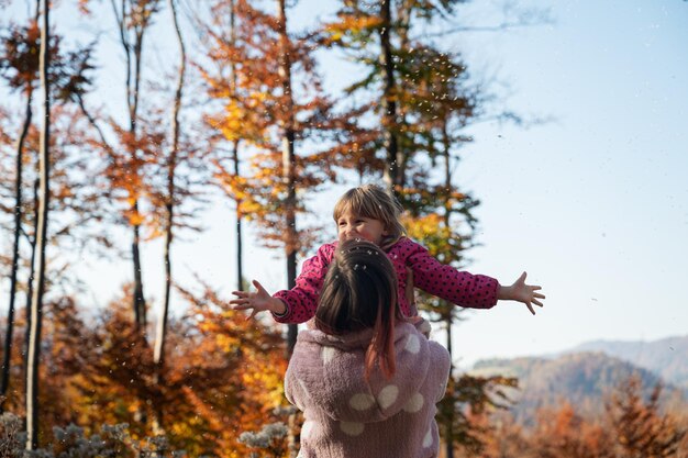 Junge Mutter, die ihre lächelnde Kleinkindtochter draußen in der schönen Herbstnatur in die Luft hebt