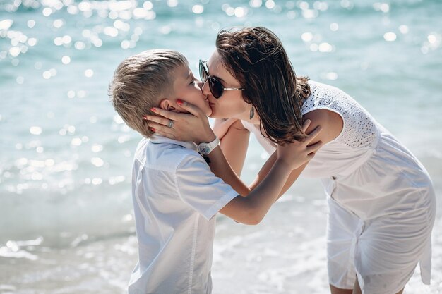 Junge Mutter, die ihren kleinen Sohn am Strand küsst. Familie in weißer Kleidung. Positive menschliche Emotionen, Gefühle, Freude. Phuket, Thailand. Schöne Meereslandschaft.