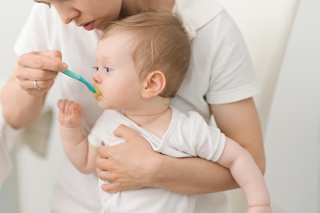 Junge Mutter, die ihren kleinen Sohn mit Fruchtpüree füttert Kleines Kind, das mit der Mutter gehalten wird, die mit einem Löffel isst