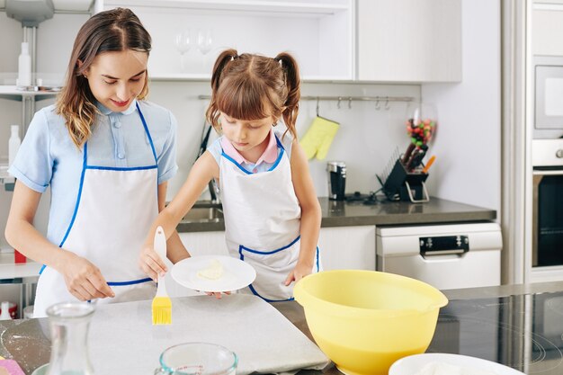 Junge Mutter, die jugendliches Mädchen mit Silikonpinsel beim Auftragen von Butter auf Pergamentpapier betrachtet