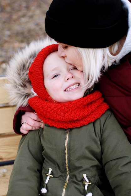 Junge Mutter, die mit ihrer süßen kleinen Tochter im Freien spricht, die in warmer Kleidung zusammensitzt