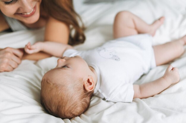 Junge Mutter, die sich mit süßem Baby auf dem Bett mit weißen Leinen-Naturtönen amüsiert, liebt Emotionen