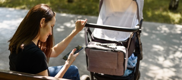 Junge Mutter, Frau mit Kinderwagen mit Smartphone in den Händen, die auf Parkbank sitzen