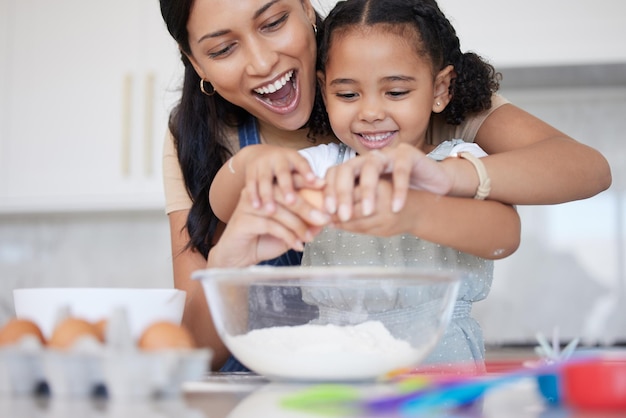 Junge Mutter genießt das Backen Bindung mit ihrer kleinen Tochter in der Küche zu Hause Little Latino Mädchen lächelt, während sie ihrer Mutter hilft, eine Mahlzeit zu Hause zu kochen Kind, das ein Ei für ein Rezept knackt