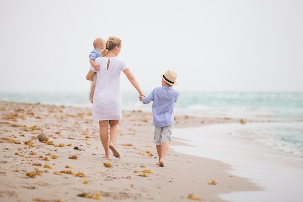 Junge Mutter im weißen Kleid, die mit ihren beiden kleinen Jungen am Meeresstrand spazieren geht