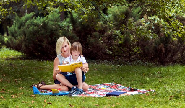Junge Mutter liest mit ihrem besonderen Sohn auf einem Teppich inmitten einer Parkwiese ein Buch