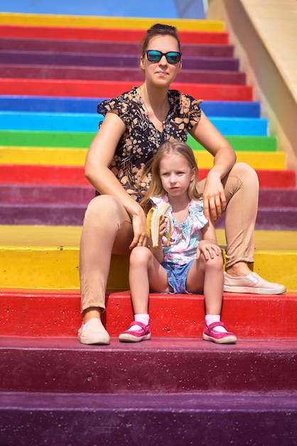 Junge Mutter mit Brille und Tochter sitzen auf einer farbigen Treppe. Kinderschutzkonzept, Muttertag