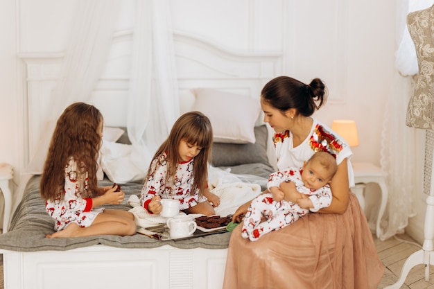 Junge Mutter mit ihrem kleinen Baby sitzt mit ihren beiden Töchtern im Schlafanzug auf dem Bett und isst Kekse mit Kakao mit Marshmallows.