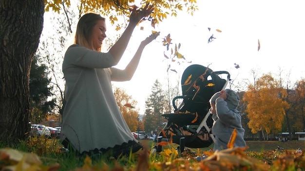 Junge Mutter mit ihrem lachenden kleinen Baby, das im Herbstpark Blätter in die Luft wirft