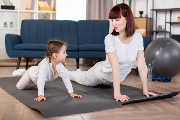 Junge Mutter mit ihrer kleinen Tochter macht Fitness-Stretching-Push-up-Übungen auf dem Boden