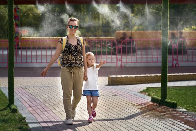 Junge Mutter mit ihrer Tochter kühlt sich bei heißem Wetter vor den Toren der Klimaanlage im Wasserpark ab. Aerosolversprühen von Wasserstrahlen, Versprühen von Kühlwasser, Klimaanlagendüse