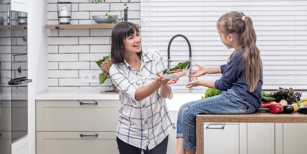 Junge Mutter mit ihrer Tochter mit viel Gemüse auf dem Hintergrund des Innenraums einer modernen hellen Küche.