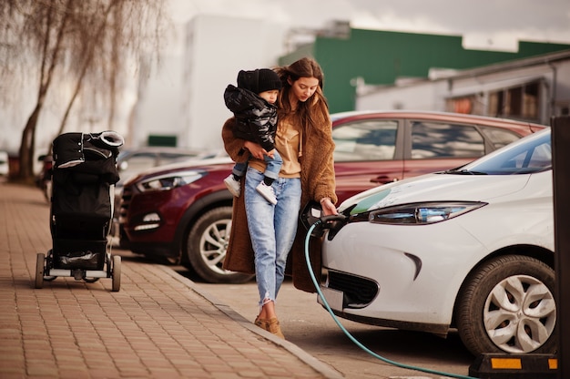 Junge Mutter mit Kind, die Elektroauto an der Elektrotankstelle auflädt.