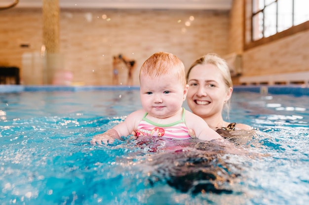 Junge Mutter Schwimmlehrerin und glückliches kleines Mädchen im Planschbecken Bringt Kleinkindern das Schwimmen bei Genießen Sie den ersten Tag des Schwimmens im Wasser Mutter hält das Kind an der Hand, das sich auf das Tauchen vorbereitet und Übungen macht