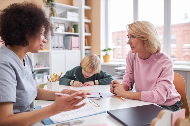 Junge Mutter spricht mit dem Kinderarzt in der Klinik mit dem Kind im Hintergrund