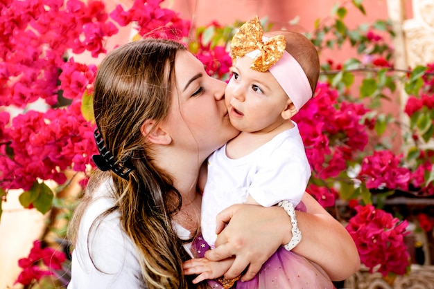 Junge Mutter umarmt und küsst ein süßes Baby vor dem Hintergrund von Blumen am Muttertag
