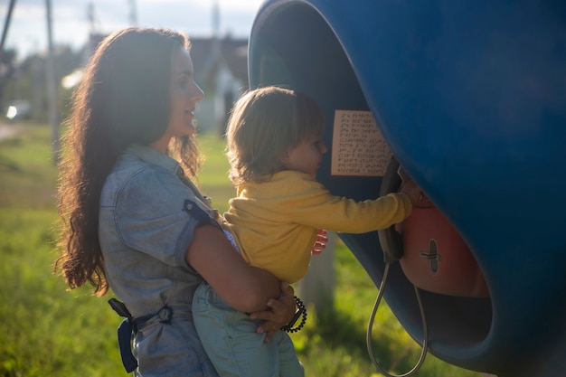 Junge Mutter und Baby rufen im Sommer über ein Festnetztelefon in der Telefonzelle im Dorf an. Wandern mit der Familie sonnigen Sommertag bei Sonnenschein. künstlerischer Schwerpunkt