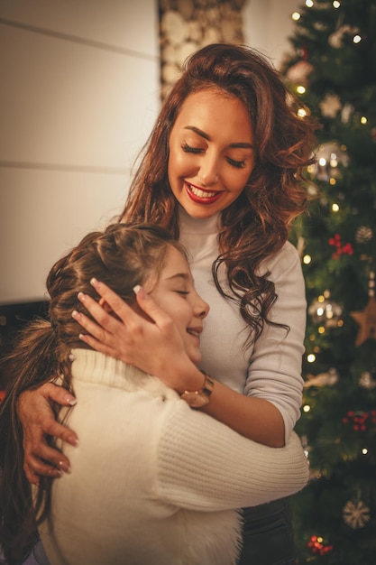 Junge Mutter und ihre süße kleine Tochter umarmen und genießen neben dem Weihnachtsbaum zu Hause.