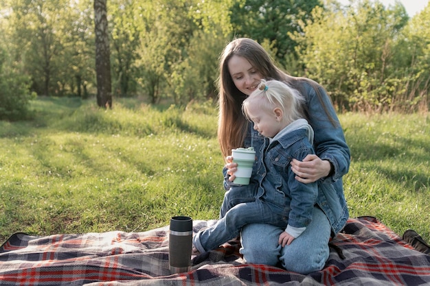 Junge Mutter und kleine Tochter auf Picknick im grünen Park am sonnigen Frühlingstag Kleines Mädchen sitzt auf dem Schoß ihrer Mutter