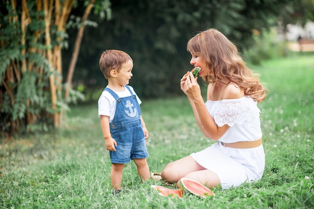 Junge Mutter und kleiner Sohn spielten auf Gras. Glückliche Mutter und Sohn sitzen im Gras und essen Wassermelone. Sommer.