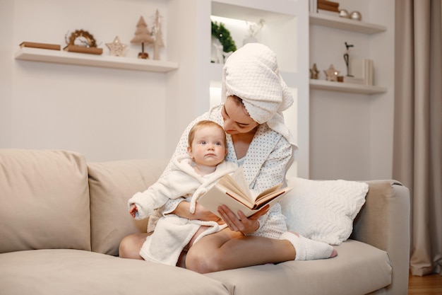 Junge Mutter und kleines Mädchen tragen Bademäntel mit Haaren in Handtücher gewickelt Frau und Mädchen sitzen auf einem Sofa Frau liest ein Buch zu ihrer Tochter