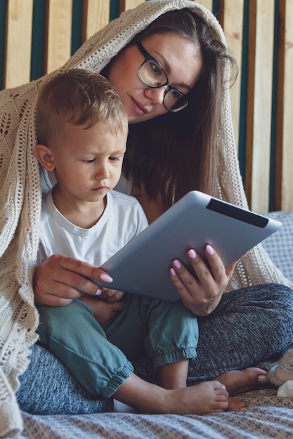 Junge Mutter und Sohn benutzen eine Tablette unter einer kuscheligen Decke im Schlafzimmer ihres Hauses