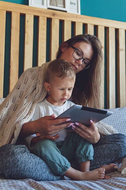 Junge Mutter und Sohn benutzen eine Tablette unter einer kuscheligen Decke im Schlafzimmer ihres Hauses