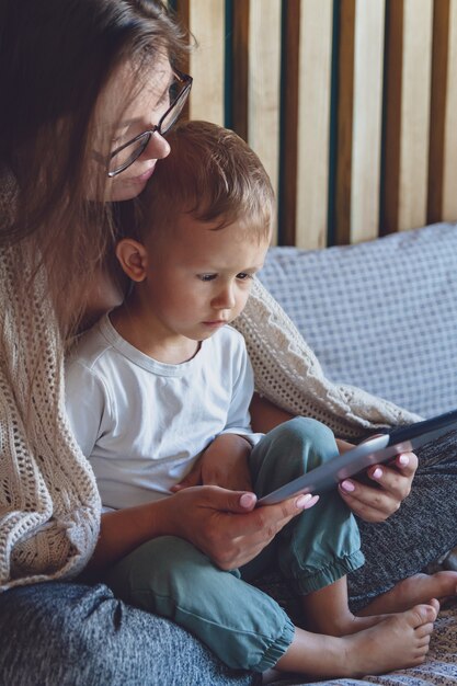 Junge Mutter und Sohn benutzen eine Tablette unter einer kuscheligen Decke im Schlafzimmer ihres Hauses