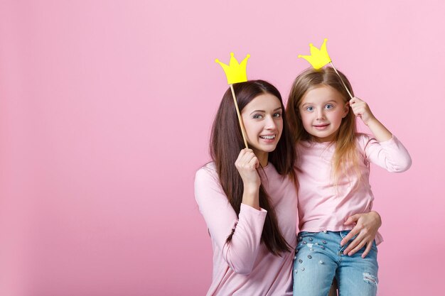 Junge Mutter und Tochter, die zusammen Spaß haben und eine gelbe Papierkrone auf rosa Hintergrund halten