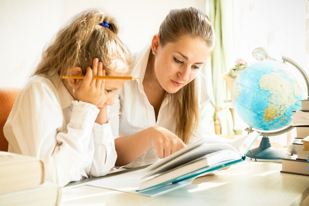 Junge Mutter und Tochter lesen Lehrbuch während der Hausaufgaben