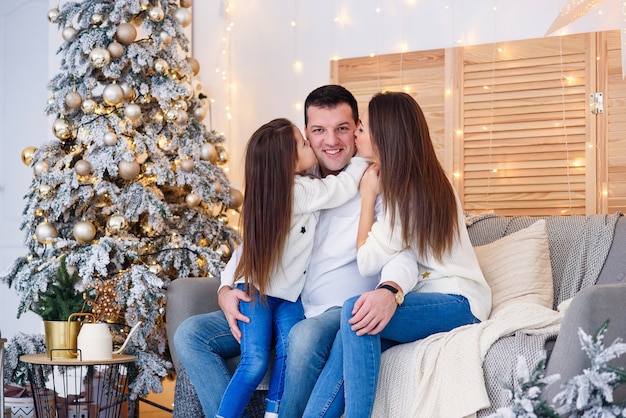 Junge Mutter, Vater und ihre kleine Tochter sitzen schön in der Nähe von Weihnachtsbaum auf Sofa.