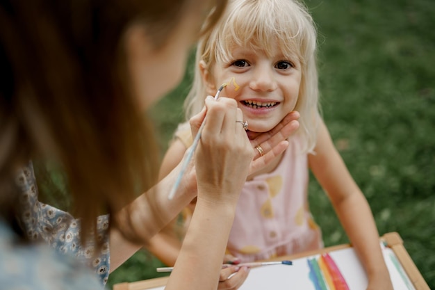 Junge Mutter zeichnet mit Farben auf ihrem Kindergesicht im Garten. Familienzeit.