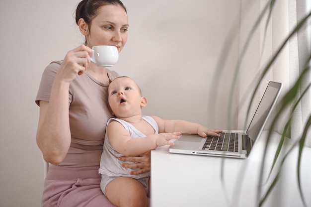 Junge Mutterarbeit, die von zu Hause mit Laptop-Computer studiert, kleines niedliches Kleinkindbaby auf Schoß.