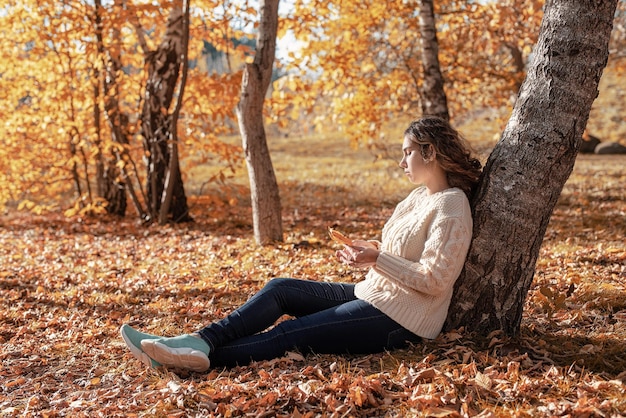 Junge nachdenkliche Frau, die am Baum im Herbstwald sitzt