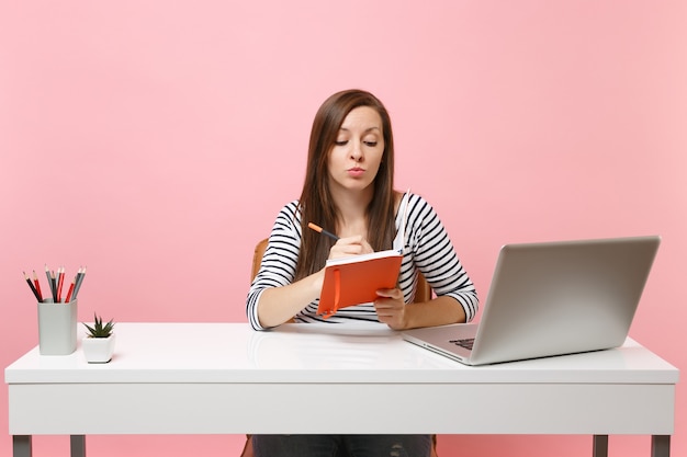 Junge nachdenkliche Frau in Freizeitkleidung, die Notizen zum Notebook schreibt und am weißen Schreibtisch mit modernem PC-Laptop arbeitet
