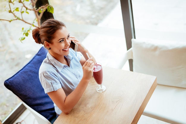 Junge nachdenkliche Geschäftsfrau in einer Pause in einem Café. Sie benutzt Smartphone und trinkt Fruchtsmoothie.