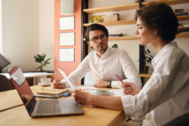 Junge nachdenkliche Geschäftskollegen diskutieren neue Projekte und sitzen mit Laptop und Papieren im modernen Büro am Tisch